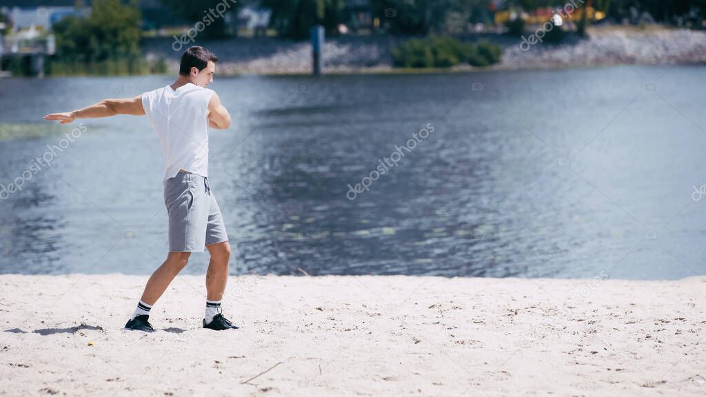 young sportsman in tank top and shorts doing body rotation near river