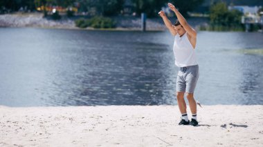 young sportsman in tank top and shorts warming up with outstretched hands near river clipart