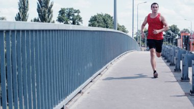 young sportsman in tank top and shorts running on bridge in summer clipart