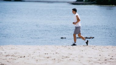 Kolsuz tişörtlü genç sporcunun nehir kenarında koşuşunun yan görüntüsü