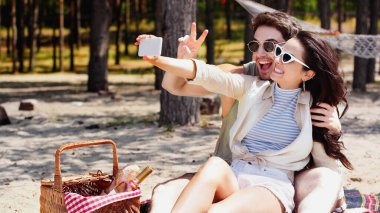 Sahilde piknik sepetinin yanında selfie çeken gülümseyen bir çift. 