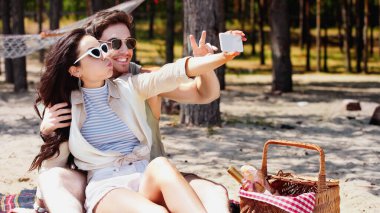Gülümseyen adam plajdaki piknik sepetinin yanında sevgilisiyle selfie çekerken barış işareti gösteriyor. 