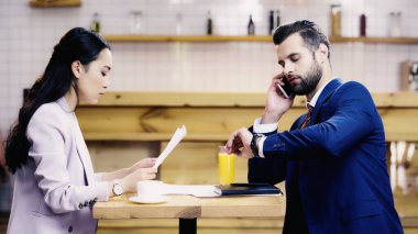 Asyalı iş kadını iş adamının yakınındaki belgelere bakıyor. Akıllı telefondan konuşuyor ve kafede saatleri kontrol ediyor.