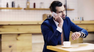 Takım elbiseli sakallı iş adamı akıllı telefondan konuşuyor ve kafede saatleri kontrol ediyor.
