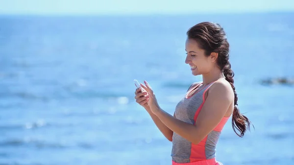 Smiling Sportswoman Using Smartphone Blurred Sea — Stock Photo, Image