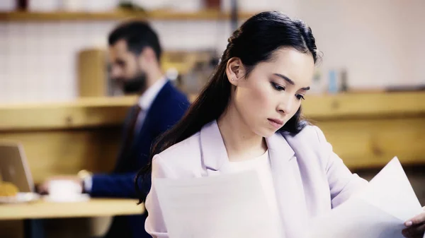 Concentrato Asiatico Businesswoman Guardando Documenti Vicino Sfocato Businessman Sfondo Caffè — Foto Stock