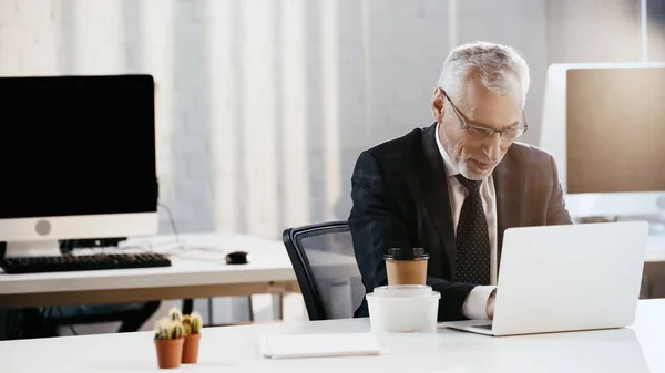 オフィスの食器やコーヒーの近くのノートパソコンを使用して眼鏡のビジネスマン — ストック写真