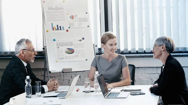 Mujer Negocios Sonriente Mirando Colega Mediana Edad Cerca Computadoras Portátiles — Foto de Stock