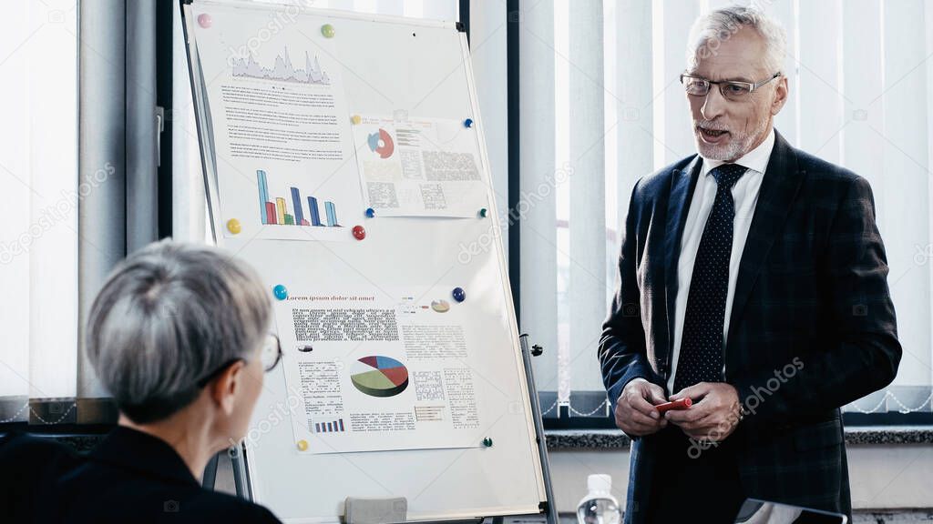 Middle aged businessman talking near blurred businesswoman and flip chart in office 