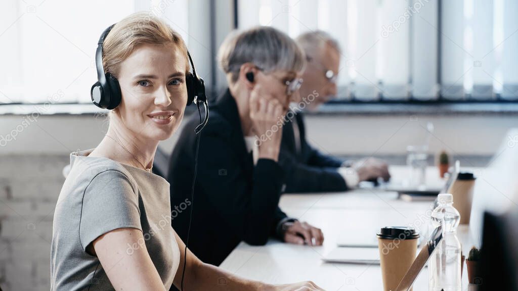 Smiling businesswoman in headset looking at camera near laptop and drinks in office 