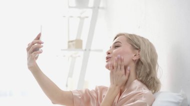 Neşeli sarışın kadın akıllı telefondan selfie çekerken hava öpücüğü gönderiyor.