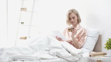 Yatakta online alışveriş yaparken elinde dijital tablet ve kredi kartı tutan sarışın kadın. 