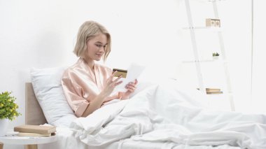 Yatakta online alışveriş yaparken elinde dijital tablet ve kredi kartı tutan gülümseyen kadın. 