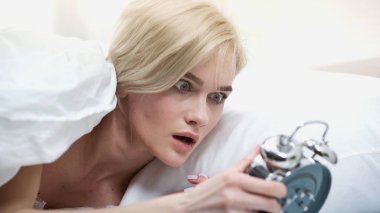 shocked young woman looking at retro alarm clock in bedroom clipart