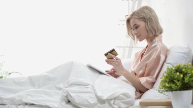 Yatakta dijital tablet ve kredi kartı tutan sarışının yan görüntüsü 