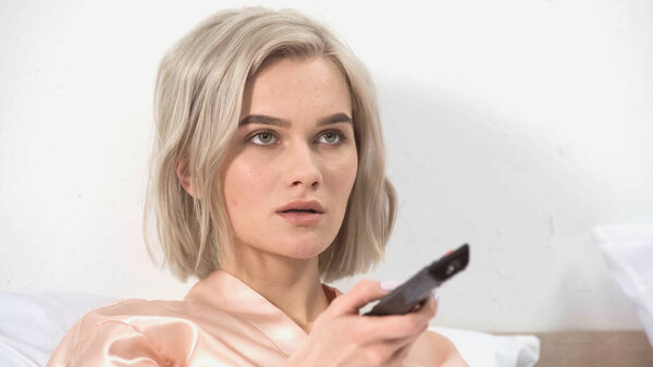 шокированная блондинка держит пульт дистанционного управления в спальне