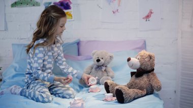 Yanında pijamayla çay fincanlarının yanında oynayan çocuk ve yatakta yumuşak oyuncaklar. 