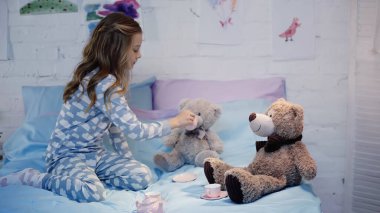 Yatakta yumuşak oyuncağın yanında pijama giymiş çocuk resmi. 