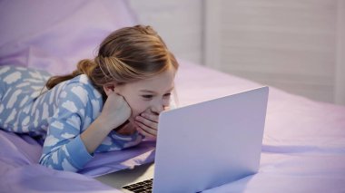 Pijamalı neşeli çocuk yatakta yatarken dizüstü bilgisayara bakıyor. 