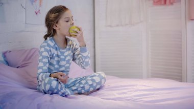 Preteen child eating apple while sitting on bed in evening 