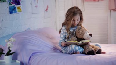 Smiling kid in pajama reading fairytale near soft toy on bed in evening  
