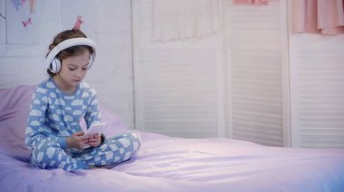 Preteen child in pajama using headphones and smartphone on bed in evening 