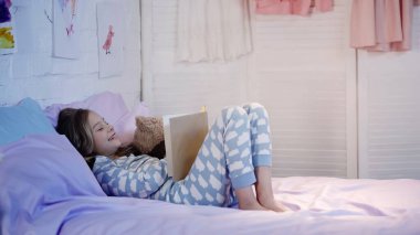 Preteen kid in pajama laughing while reading book near teddy bear on bed 