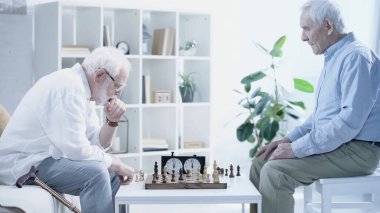 side view of thoughtful man playing chess with senior friend in living room