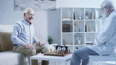 cheerful senior man looking at friend sitting near chessboard in living room