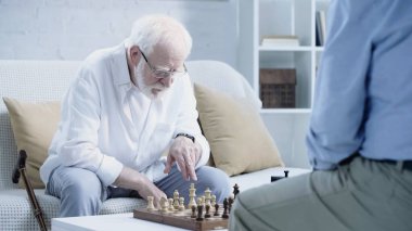 senior man in eyeglasses pointing at chessboard near blurred friend