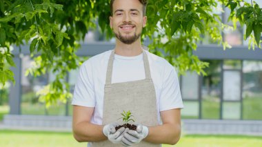 Smiling gardener in gloves holding plant in soil in garden 