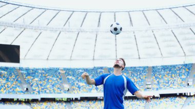 Mavi tişörtlü sakallı futbolcu. Stadyumun üstünde kafası olan zıplayan top. 