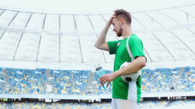 Yeşil tişörtlü genç futbolcunun saçını düzeltirken elinde top tutarken ve spor şişesi tutarken yandan görünüşü