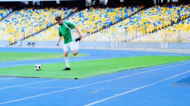 full length of bearded football player in uniform warming up near ball at stadium
