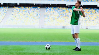 full length of bearded football player in green uniform warming up near ball at stadium