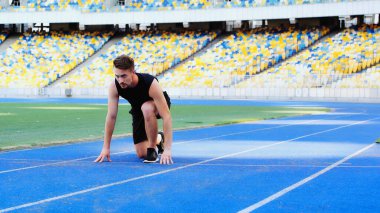 Spor giyimli ve spor ayakkabılı sakallı bir adam stadyumda koşmadan önce başlangıç pozisyonunda duruyor. 