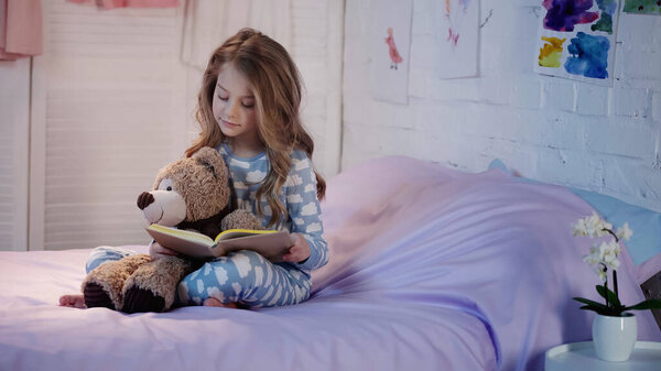 Preteen child in pajama reading book near teddy bear in bedroom 