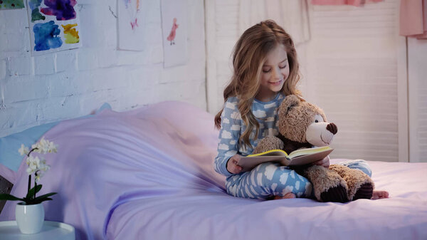 Smiling preteen child in pajama reading book near soft toy on bed in evening 