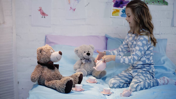 Side view of kid in pajama pouring tea near teddy bears on bed 