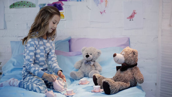 Side view of girl in pajama pouring tea near cups and soft toys on bed 