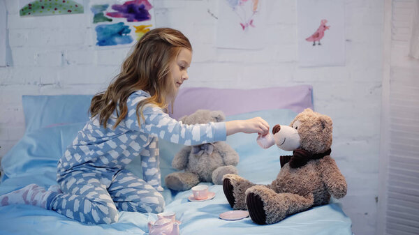 Side view of girl in pajama holding cup near teddy bear on bed 