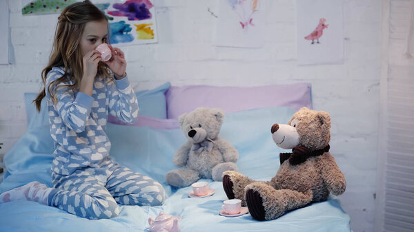 Preteen child drinking tea near soft toys on bed in evening 