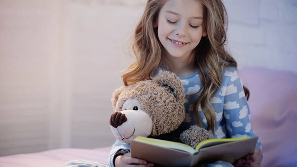 Cheerful preteen kid holding teddy bear and reading book on bed 