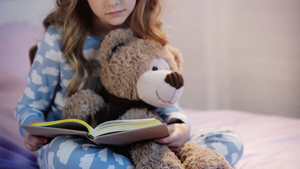 Cropped view of preteen child reading book near blurred teddy bear on bed 