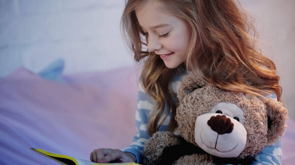 Cheerful preteen child in pajama reading book near soft toy in blurred bedroom 