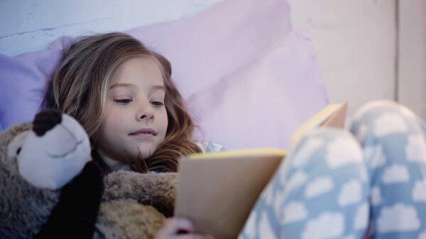 Preteen kid reading book while lying near teddy bear on bed 