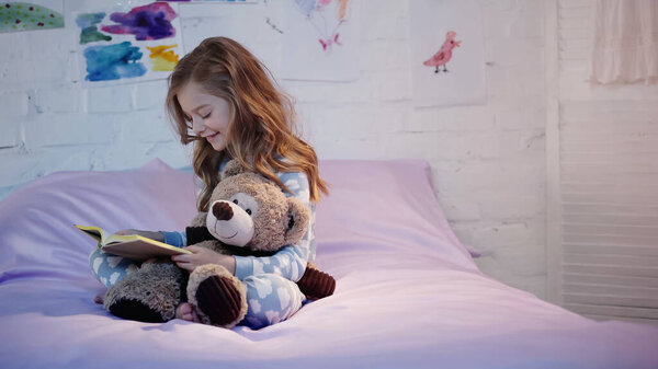 Happy preteen kid reading book and hugging soft toy on bed in bedroom 