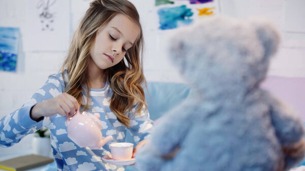 Preteen child in pajama pouring tea near blurred soft toy in bedroom 