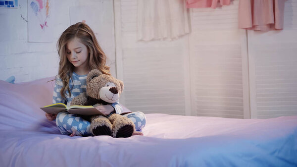 Child in pajama reading fairytale near teddy bear on bed in evening  