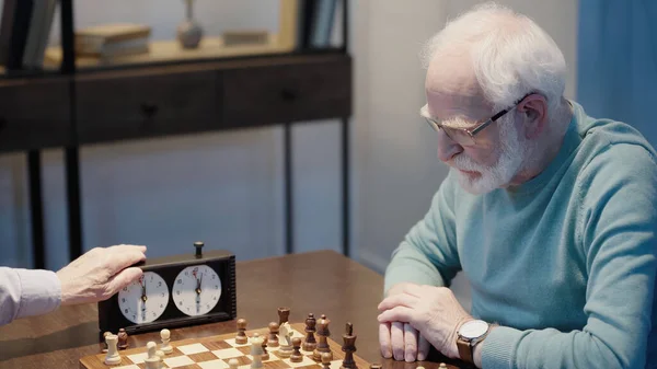 grey haired man thinking near chessboard and friend fixing time on chess clock